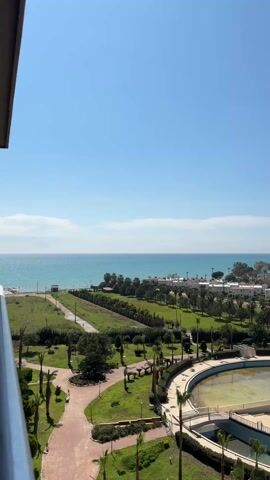 The video provides a high-angle, wide-shot view of a coastal landscape on a sunny day. The blue sky is mostly clear, with a few wisps of white clouds. The turquoise sea stretches to the horizon, meeting the sky in a hazy line. In the midground, there's a green park area featuring a mixture of grass, trees, and palm trees. Walking paths meander through the park, constructed of reddish-brown material. A circular, empty water feature dominates the foreground of the park. The structure is made of concrete. Some white buildings are nestled amidst the greenery near the coastline. A few birds fly in the sky. A portion of a building, perhaps a balcony, is visible in the far left of the frame.