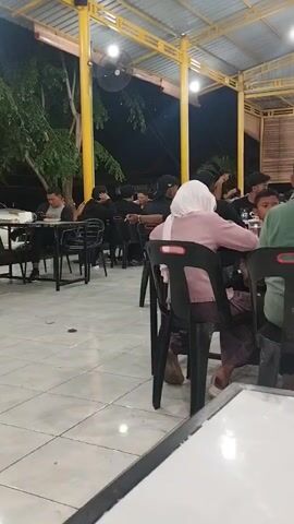 The video shows a few people eating at an outdoor restaurant. Several tables are occupied by groups of people of various ages. Some people wear head coverings. The building has a corrugated metal roof supported by yellow posts, and a few fans are mounted overhead. It is nighttime.