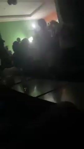 The video shows a dimly lit indoor space with several people. The room has a green wall and a white ceiling, with a fan visible. Some individuals appear to be standing, while others are seated. There's a table with a bottle on it. A person is recording the scene from a low angle, potentially while sitting. The lighting is poor, making details difficult to discern.