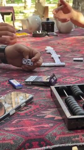 The video shows a close-up of a table where a group of people are playing dominoes. The table is covered with a red, patterned cloth. In the foreground, a hand holds a domino tile with two dots on one end and one dot on the other. Next to the dominoes are a cigarette pack, a lighter, and a smartphone. To the right of the frame is an abacus with black beads. In the background, other hands are visible playing dominoes, and several teapots are arranged on the table. The setting appears to be outdoors, with trees and sunlight visible in the background. The focus is on the game and the objects on the table.