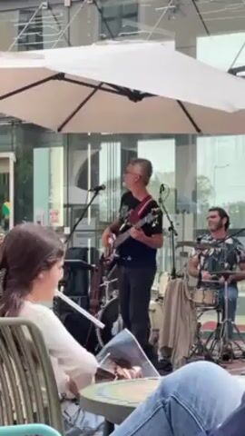 A band performs outdoors under a white patio umbrella. The band consists of a bass player/singer and a drummer. The bass player is in the center wearing a black t-shirt and jeans, playing a red and brown bass. A mic stand stands in front of him. The drummer is seated on the right wearing a floral shirt. The drums are a small kit. In the foreground on the left, a young woman with long brown hair sits at a table, reading a magazine or book. People's legs and blue jeans are visible across the bottom of the frame. The background features a modern building with glass walls.