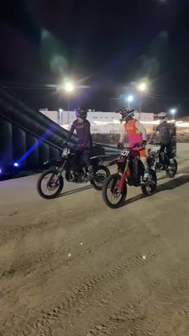 A nighttime outdoor event features three dirt bike riders in full gear. The rider in the foreground on the left wears purple and rides a black dirt bike. To their right, a rider in pink and orange with the number 27 on their bike rides a red dirt bike. Behind them, another rider in a grey shirt and helmet on a black dirt bike is partially visible. In the background, a large dark inflatable ramp structure is positioned to the left. Beyond that, spectators are visible, along with lights and trees. The ground is dirt with visible tire tracks. The riders appear to be preparing for or participating in a race or stunt event.