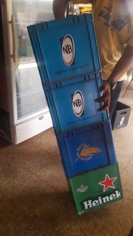 A person in a yellow shirt is seen struggling to hold a stack of beer crates. The crates are stacked unevenly and appear unsteady. There are three blue crates with the logo "NB Nigerian Breweries Plc." on top of a green Heineken crate. The person attempts to adjust the stack, but it remains tilted. The crates appear to contain beer bottles. The scene is inside a building with a tiled floor and the edge of a refrigerator visible in the background.