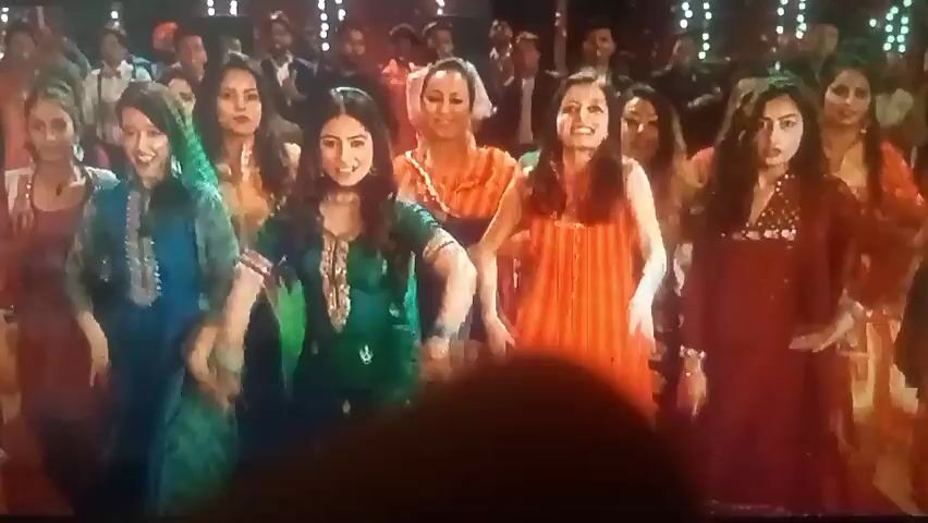 The video shows a group of women in colorful traditional Indian clothing dancing at an event. They are surrounded by a crowd of people, some of whom are also dancing or watching. The women are smiling and appear to be enjoying themselves. A young man in a green and white striped sweater stands out in the crowd, looking serious and singing. The background is decorated with lights.