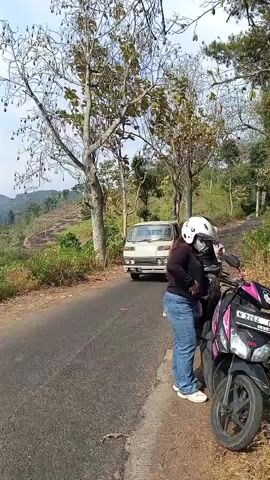 The video opens with a view of a rural road. A white pickup truck is seen approaching from the left, and a motorcycle with two people is parked on the right side of the road, with the person in the foreground wearing a white helmet and blue jeans, and the person behind wearing a black shirt and also a helmet. As the truck passes, the camera pans to follow the motorcycle. The truck continues down the road and then the camera focuses on the two people on the motorcycle. The person in the foreground adjusts their helmet, and then the person behind them starts the motorcycle. The video ends as the motorcycle begins to move. The environment appears to be a hilly, vegetated area with trees and dry grass.