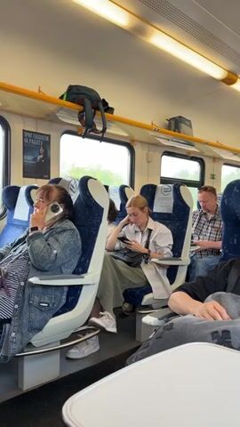 This is a video shot from inside a train carriage, looking forward and slightly to the side. Several passengers are visible, mostly seated and engaged with their phones or looking out the window. The seats are blue and white, with overhead luggage racks holding bags. The interior is lit by bright fluorescent lights. Outside the window, greenery and trees are visible, with a white wall running alongside the tracks. In the distance, a red object, possibly part of a train or structure, can be seen. The overall atmosphere is that of a typical train journey.
