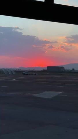 The video shows a red sunset viewed from inside a building, possibly an airport terminal, as the foreground shows a tarmac with markings and a white work truck with a trailer parked nearby. In the background, mountains are silhouetted against the vibrant red and pink sky with scattered clouds. The framing of the video includes part of the building's structure at the top, creating a rectangular border. The scene is still, with no discernible movement except for the subtle shift in color as the sun sets.
