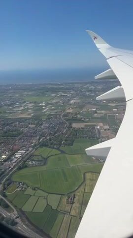 The view from an airplane window shows a white wing with several flaps against a landscape of patchwork green and brown fields and a small town. In the distance, a thin strip of land separates the fields from the blue ocean under a clear blue sky. A highway is visible at the bottom of the frame. The airplane belongs to Qatar Airways, as seen by the logo on the wing.