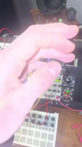 The video shows a hand with tattoos manipulating the controls of a modular synthesizer. The synthesizer is black with various modules connected by colorful patch cables. The hand adjusts knobs and presses buttons on the Quad VCA module. In the background, a wooden shelf holds a Genelec speaker, figurines, and other small objects. A keyboard is visible at the bottom of the frame.