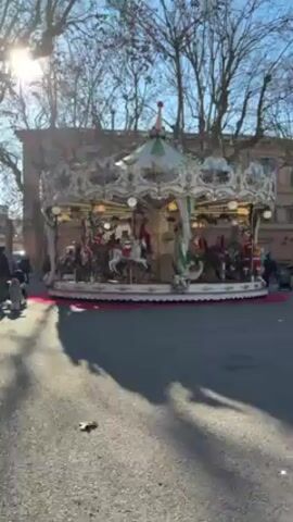 The video shows a carousel spinning in an outdoor setting. The carousel is adorned with lights and detailed carvings and paintings. The platform beneath it is covered in a red material. Trees surround the carousel and a building is visible in the background. The sun is shining brightly, casting shadows on the ground. People can be seen walking in the background. The camera is mostly static.
