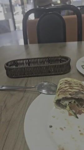 The video shows a close-up of a table in a restaurant. On the table, there is a plate with a partially eaten wrap, a spoon, and a small woven basket. In the background, two people wearing hats are seated at a table, facing away from the camera. Through the large windows behind them, a street scene is visible, suggesting an urban setting. The overall lighting indicates it's daytime, possibly late morning or early afternoon. The atmosphere appears casual, consistent with a mealtime at a cafe or restaurant. The blurriness of the background and the focus on the food suggest the recording is primarily about the meal or the dining experience. No discernible text is visible in the video. The location is identified as Karakol City, Kyrgyzstan.