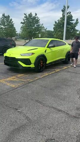 The video shows a bright green Lamborghini Urus parked in a parking lot. The car is in the foreground, taking up most of the frame. It has black accents, including the wheels and front grill, with orange brake calipers. A person walks past the car on the right side of the frame. The parking lot is marked with yellow lines. In the background, there are trees, a light pole, and other parked vehicles. The sky is partly cloudy. The camera slowly zooms in on the front of the car.