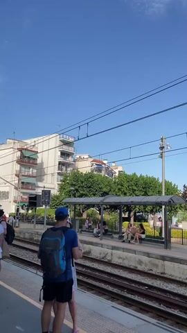 A train station platform is shown under a clear blue sky. On the platform, several people are sitting and standing under a covered waiting area. A man wearing a blue t-shirt and a backpack stands in the foreground, facing away from the camera, with another person next to him. Across the tracks, a sign with the words "SANT'POL de FURS" is visible, with trees and the sea in the background. A digital display board showing a green light is also visible. The scene appears to be a warm, sunny day.