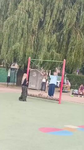 Three young people are playing on a playground. A girl with long blonde hair stands to the left, facing away from the camera, wearing camouflage pants and a black top. In the center, a girl with long brown hair is hanging from a horizontal bar with her arms. She is wearing a white crop top and dark pants. To the right, a person with blonde hair is also hanging from the bar, wearing a light blue long-sleeved shirt and dark pants. They are positioned on a set of parallel bars suspended by ropes from a larger horizontal bar. The playground surface is green, with colorful painted shapes. Behind them, a large weeping willow tree with long, drooping branches dominates the background. To the left of the tree, a gray portable toilet with a white logo is visible. Further in the background, there are buildings, fences, and other playground equipment, including a swing set with people on it. The overall scene suggests a casual outdoor activity on a cloudy day.