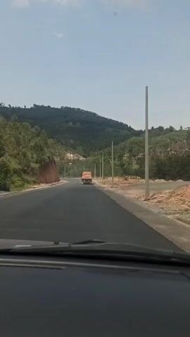A car drives on a paved road, approaching an orange truck in the distance. The road is surrounded by hills with trees. There are utility poles alongside the road. The sky is blue.
