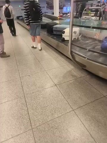 The video shows a luggage carousel at an airport. Several pieces of luggage are visible on the conveyor belt, including a light blue suitcase with pink trim, a white suitcase with a black handle, and several black suitcases of various sizes. In the background, several people are standing near the carousel, presumably waiting for their luggage. Some of them appear to be looking at the conveyor belt, while others are talking to each other. The overall scene is that of a busy airport baggage claim area. There are signs visible in the background, one of which reads "2". The floor is tiled and appears to be speckled grey. The lighting suggests it is daytime, likely indoors as is typical for an airport. There are no obvious weather cues, as the scene is entirely within the airport. The emotions of the people are not clearly discernible from the video. The text visible includes "2" on a pillar and some indistinct text on screens in the background. The location is Kastrup, Denmark.