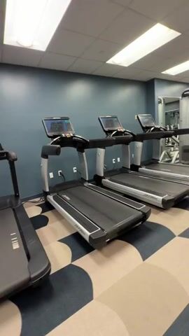 The video shows an interior view of a gym. In the foreground, a row of treadmills is visible, with their consoles illuminated. The treadmills are sleek and modern. Behind the treadmills, the gym continues with weight machines and mirrors lining the walls. The walls are painted a muted blue, and the floor features a distinctive geometric pattern of beige and dark blue squares. Fluorescent lights are installed in the ceiling, providing ample illumination for the space. There are no people visible in the video. The camera pans slowly from left to right, offering a sweeping view of the gym's layout and equipment.