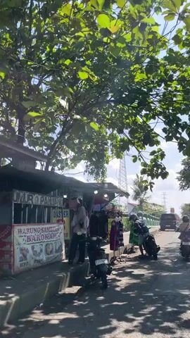 The video shows a street scene with a food stall on the left. The stall has signs in Indonesian, one reading "WARUNG SAM... BAROKAH" and another with images of chicken. Several people are standing at the stall. There are motorcycles parked in front of the stall. The street is paved and appears to be a public road. Trees with green foliage overhang the scene, providing shade. In the background, there is a transmission tower and a vehicle on the road. The sun is shining, creating shadows. The overall impression is of a busy roadside market or eatery.
