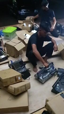 This video depicts a scene of people unpacking and assembling equipment, likely for an event or setup.

In the first segment, two men are seen on the floor surrounded by cardboard boxes and electronic equipment. One man, wearing a black t-shirt, is crouching and appears to be examining or handling a piece of hardware. The other man, also in a black t-shirt, is standing and opening a box. The setting seems to be indoors or under a covered area, with lighting suggesting it could be evening or a dimly lit space. There are stacks of boxes, some open, with electronic components visible. One box has a green lid.

The second segment continues with a similar scene. The man in the black t-shirt is still crouched, focused on the equipment. More electronic devices, possibly computer parts or networking equipment, are scattered around. A tarp or cloth is visible overhead, providing cover. The lighting is a bit brighter here, with a visible light source illuminating the area. The context of Mideuenjok, Indonesia, suggests this could be related to a local event or business setup.

The third and fourth segments show a transition to a different scene, with a more dynamic, perhaps handheld camera movement, making it difficult to discern clear details. The final segment shows a man in a teal shirt and a cap, wearing gloves, working on a large, grey panel against a dark background. Metal structures are visible overhead, suggesting an industrial or event setup environment. The man is leaning against the panel, possibly installing or adjusting it. The overall impression is one of labor and preparation for an event or construction.