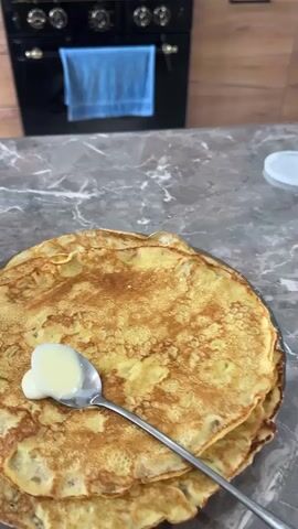 A spoon is used to spread a creamy white substance on top of a stack of golden brown crepes. The crepes are arranged on a gray speckled countertop, with a portion of a black gas stove visible in the background, adorned with a blue dish towel hanging from the oven door.