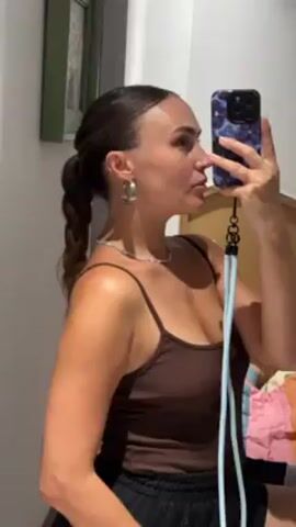A woman takes a selfie. She faces to the side in the frame, her head in profile as she holds a cell phone up to capture her reflection in a mirror. She wears a brown tank top, black shorts, hoop earrings, and a silver choker necklace. Her hair is pulled back in a braided ponytail. The phone has a blue patterned case and a light blue cord attached to it. A portion of a wooden headboard is visible behind the woman, along with a framed picture on the wall to the left.