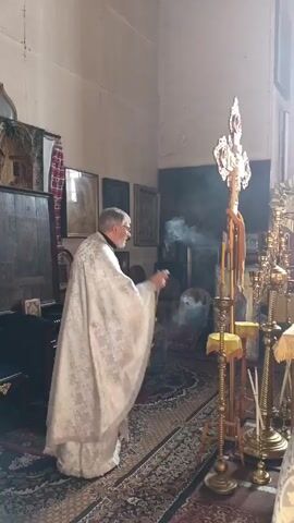 The video shows a priest in a white robe standing in a room with religious objects, swinging a censor that emits smoke. The priest is an older man with a gray beard. He stands in front of a table on which there is a gold cross, candlesticks, and other ornate objects. There are also icons hanging on the walls. The room is carpeted with a Persian-style rug. Another person, also in a white robe, is partially visible to the right. The light in the room is soft, suggesting a natural light source from a window.
