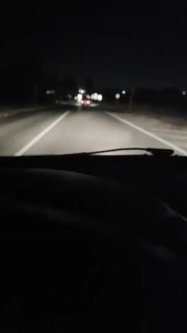 A first-person perspective video captures a night drive on a rural road. The headlights of the vehicle illuminate the asphalt, casting a glow on the white lane markers. In the distance, other vehicles can be seen, their taillights and headlights faintly visible. The environment appears dark with trees lining the sides of the road, and some signs are visible along the route. The video is short and appears to be a continuous shot.