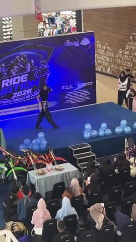 The video shows a young person with long dark hair wearing a black crop top and dark pants with a white sash dancing on a blue stage. The stage is decorated with blue balloons and has a backdrop with the text "RIDE NATION 2026" and logos for "CITIMALL DUMAI X IMI DUMAI". There are also two dirt bikes on display to the left of the stage, and a table with some items on it. An audience is seated in black chairs facing the stage, many of whom are holding up phones to record. The event appears to be taking place indoors, possibly in a shopping mall, with the background showing store displays. Several people are visible standing on a platform to the right of the stage, observing the performance. The lighting is primarily blue, coming from the stage backdrop.
