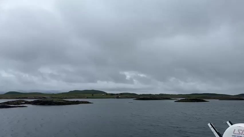 This is a video taken from a moving vessel on a body of water. The sky is heavily overcast with dark, grey clouds. In the distance, there are several small, rocky islands covered with green vegetation. A few small buildings, possibly houses, are visible on some of the islands. The water is dark and choppy, with a white wake trailing behind the vessel on the left side of the frame. The overall impression is of a dreary, possibly cold, maritime environment.