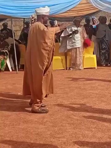 An outdoor event is taking place under a blue and white tent, with people gathered on a reddish-brown dirt ground. The central figure is a dark-skinned man in traditional attire, including a light-colored cap, a tan agbada, and dark shoes. He is holding a white smartphone horizontally with both hands, appearing to record or photograph something in front of him. His back is to the camera for most of the video, but he briefly turns his head and body to his left. Several other individuals, also dressed in traditional clothing, are visible in the background, sitting on chairs or standing. Some yellow chairs are arranged in rows.