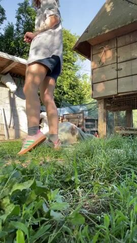 A daytime video clip, filmed in Danilovgrad, Montenegro, shows a woman walking in a grassy outdoor area. She is wearing a light-colored, long-sleeved shirt, denim shorts, and sneakers. The setting includes a wooden chicken coop on the right side of the frame and other rustic wooden structures on the left, with green trees visible in the background under a clear sky. The woman walks from left to right, then turns to face the camera, offering a slight smile before standing still. The camera is positioned low to the ground, with lush green grass filling the foreground.
