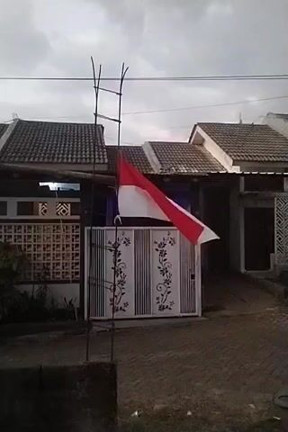 A red and white Indonesian flag is mounted on a makeshift flagpole constructed from metal poles and bamboo. The flagpole is positioned in front of a residential property with a white metal gate decorated with floral patterns. The houses in the background have tiled roofs. The sky is overcast, suggesting a cloudy day. The flag appears to be blowing in the wind.