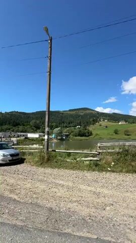 The video shows a view of a rural landscape on a sunny day. In the foreground, there is a dirt road and a silver station wagon parked. Further back, there is a red car, a wooden building with a sign that says "Артур", and a body of water. In the background, there are green hills covered with trees and a clear blue sky. The camera pans to the right, revealing more of the landscape and a wooden fence.