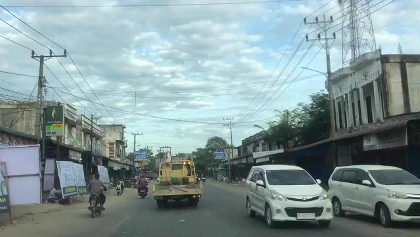 The video shows a view from a vehicle driving down a paved road in a town. The road is lined with buildings on both sides that appear to be shops or small businesses. There are many power lines and utility poles overhead. Several motorcycles are visible on the road, some carrying passengers. A yellow truck is in front of the vehicle. The sky is cloudy.