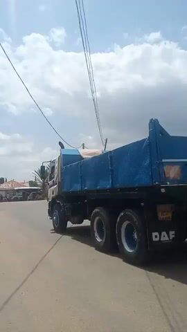 The video captures a bright, sunny day in Baccure Kofare, Nigeria. The scene is a dusty, paved road lined with buildings and vegetation. In the foreground, a young child in a dark dress walks away from the camera. The primary focus is a large, blue DAF dump truck moving down the road. The truck is empty, with its bed tilted upwards. The sky is filled with scattered clouds, indicating fair weather. Overhead, thick power lines stretch across the frame, supported by utility poles. The buildings in the background appear to be modest homes with corrugated metal roofs. The overall atmosphere is that of a typical day in a rural or semi-urban Nigerian setting. The visible text on the back of the truck is "DAF" and a license plate that reads "ICZ 7A69".