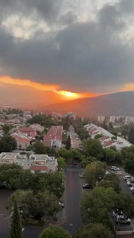 The video shows an aerial view of a residential area at sunset. The sky is filled with dramatic orange and gray clouds, with the sun peeking through the mountains in the distance. The town below consists of houses with red tile roofs, interspersed with trees. A street runs through the middle of the frame, lined with parked cars. The overall mood is serene and picturesque.