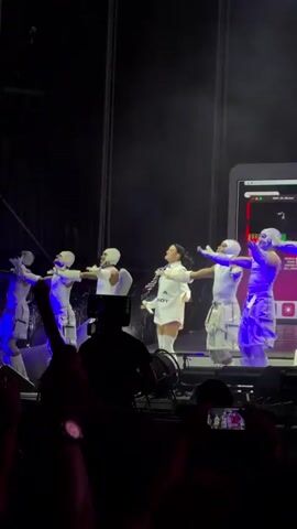 A group of performers are on a stage, illuminated by blue lights. A female performer in the center wears a white outfit with text on it. Five male performers in white outfits surround her, performing synchronized dance moves. A large screen or structure resembling a computer monitor is visible in the background. The stage appears to be outdoors, with a dark sky and visible support structures. In the foreground, audience members are visible, with some holding up cameras and phones.