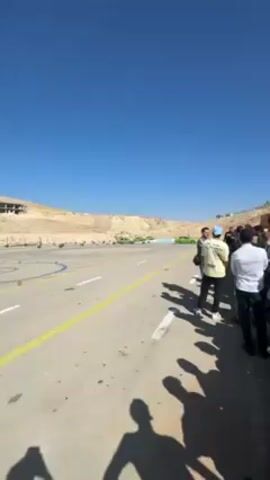 The video shows a green car drifting around a course on a sunny day. The car is a small sedan with its roof cut off, and there are two people inside. The course is marked with cones and barriers, and there is a crowd of people watching. The environment looks like an area in a desert or rocky area, under a cloudless blue sky.