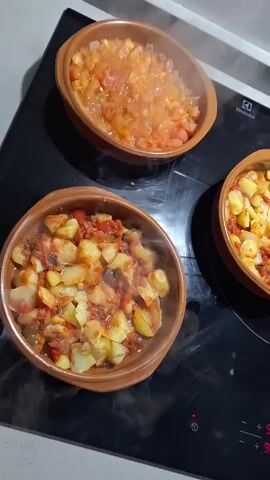 Here's a plain-text description of the video:

The video shows a black stovetop with three brown earthenware pots on it. Two of the pots contain a mixture of diced potatoes and a red sauce. Steam rises from these pots. The third pot contains a lighter red mixture of what appears to be diced tomatoes and onions. The pots are arranged in a triangular pattern on the stovetop. The camera moves slightly to provide different angles of the food. The brand "Electrolux" is visible on the stovetop.
