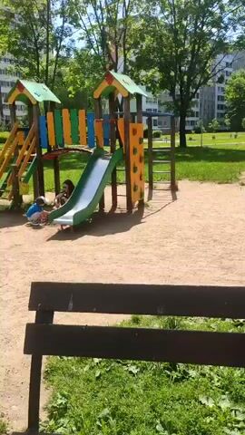 The video shows a playground on a sunny day. The playground has a slide, climbing wall, swings, and a seesaw. There are several people in the playground, including children and adults. A young girl in a pink dress is playing on the seesaw. Another child is going up the stairs of the playground structure. An adult male is walking near the playground structure. In the background, there are trees, buildings, and a paved path.