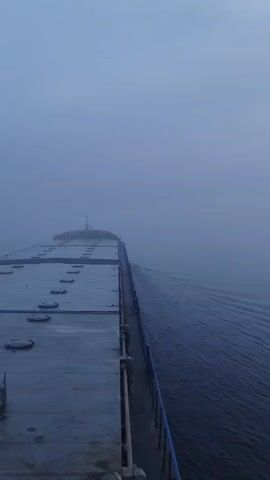 The video shows the deck of a large, flat-topped vessel traveling through water. The vessel's deck is grey and covered in round hatches. A blue railing runs along the edge of the deck. The vessel's wake creates ripples in the water. The sky is overcast and foggy, obscuring the horizon. As the camera pans, a blurred sun becomes visible through the fog.