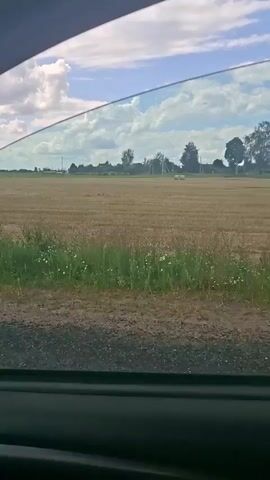 The video is an exterior shot looking out from inside a vehicle. The scene depicts a rural landscape with a harvested field of golden-brown stubble. Several large, round hay bales are visible scattered across the field. A line of green grass runs along the edge of the field nearest the viewer, bordering a gravelly area. In the distance, trees and some indistinct structures are visible under a bright, partly cloudy sky. The camera appears to be moving, indicated by the slight blur and the changing perspective of the hay bales. A reflective, orange and black pole is visible on the right side of the frame.