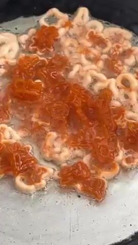 A close-up shot depicts various white, doughy shapes—letters and stars—submerged in hot oil, accompanied by a clump of vibrant orange sauce in the center. The mixture sizzles on a grey-silver cooking surface, indicating frying. The video focuses on the cooking process without showing a person or their interactions.