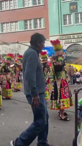 The video shows a parade taking place on a street lined with buildings and onlookers. The main focus is on a group of people dressed in elaborate, colorful costumes, likely representing a specific cultural or celebratory theme. Many of the costumes feature tall, decorated headdresses.

Alongside the parade participants, there are bystanders, including some individuals who appear to be throwing confetti or foam, creating a celebratory atmosphere. The crowd is watching the parade. 

The buildings surrounding the street range in color, with some being brick and others painted green. There are windows visible on each building. The video is taken from a perspective that follows the parade procession.