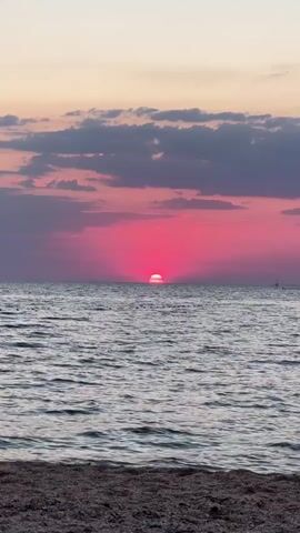 This media file captures a sunset over the sea. The sun, a bright pink orb, is low on the horizon, casting a vibrant pink glow across the sky and reflecting on the rippling water. Dark grey clouds with pinkish hues are scattered across the upper portion of the sky, adding depth and texture to the scene. The foreground shows a beach covered in small pebbles or gravel. The overall atmosphere is serene and picturesque, evoking a sense of tranquility. The time of day is clearly sunset. There are no visible people or man-made objects apart from a faint, distant silhouette on the right horizon which could be a distant structure or buoy. The location is likely a coastal area, given the presence of the sea and beach. The weather appears calm, with gentle waves on the water. There is no visible text in the media file.