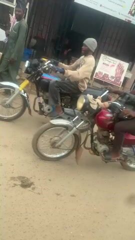 The video shows a street scene in front of a row of small businesses.  The street is paved but looks worn.  A motorcycle is parked on the side of the road. Another motorcycle drives past.  Several people are sitting in white plastic chairs in front of one of the businesses, which has a food display case labeled "DEKS Kitchen".  Two girls in school uniforms walk past in the foreground. There is also a generator in front of another shop, which has a sign that says "Teenara Nails Bar".