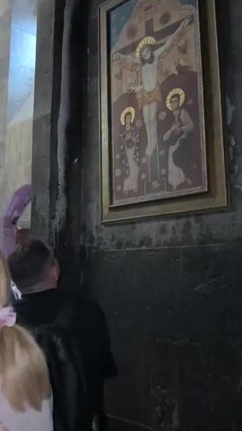 The video shows the interior of a stone church, likely an Armenian church, with several people present. The camera pans across the scene, capturing religious artwork and architectural features. 

At the beginning, a man is seen looking up at a painting of Jesus Christ on the cross, with Mary and John the Baptist depicted to his sides. This painting is framed in gold and hangs on a dark stone wall. To the left of this painting, a purple fabric hangs, possibly part of a liturgical banner.

The camera then moves to show other parts of the church interior. There are stone arches and vaulted ceilings, illuminated by chandeliers and natural light from windows. Another religious painting, depicting a saint in orange robes, is visible on a wall. In the background, an altar area with more religious imagery and candles can be seen. Several individuals are walking or standing in the church, some looking at the artwork or conversing. The overall atmosphere appears to be one of reverence and tourism.