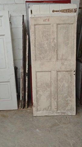 The video shows a storage room with several old doors leaning against a white brick wall. Initially, the camera focuses on two white doors, one with a "PE8" label on the top, showing signs of wear and a scratched surface. Next to it is another white door, marked "HK3". The camera pans to reveal a stack of unpainted wooden doors. Some of the doors have old-fashioned metal latches attached. In the background, there is a large container encased in a metal grid. The setting appears to be a warehouse or storage facility.