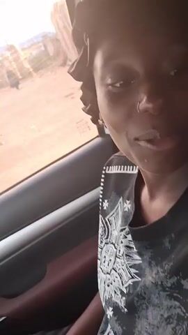 A woman is sitting in a car. She's wearing a black shirt with white decorative embellishments, a black bonnet, and a small nose ring. She smiles at the camera and her teeth are white. Light is reflecting on her face. Outside the window is visible, and looks like a semi-rural, sparsely populated area.