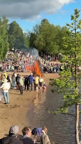 The video shows a large bonfire on a sandy beach. Many people are gathered around the bonfire, some sitting and some standing. The bonfire is very tall and made of logs. Smoke is rising from the fire. Some people are standing near the water's edge, which is visible in the foreground. The overall scene appears to be a gathering or celebration.